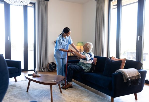 biracial female health worker helping caucasian senior man to get up from the couch at home. medical care and retirement senior lifestyle concept