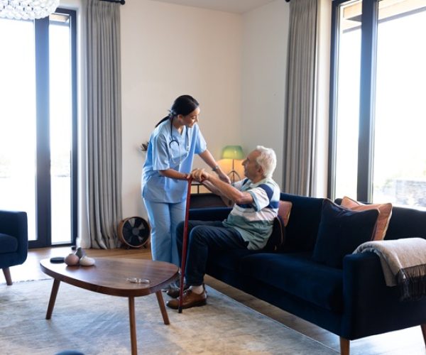 biracial female health worker helping caucasian senior man to get up from the couch at home. medical care and retirement senior lifestyle concept