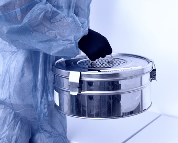 person standing in laboratory holding autoclave sterilizer metal box wearing protective uniform. blue toned