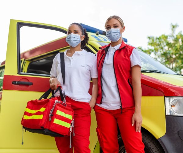 two female paramedics standing by the ambulance. two emergency nurses with face mask. paramedic carrying a medical trauma bag
